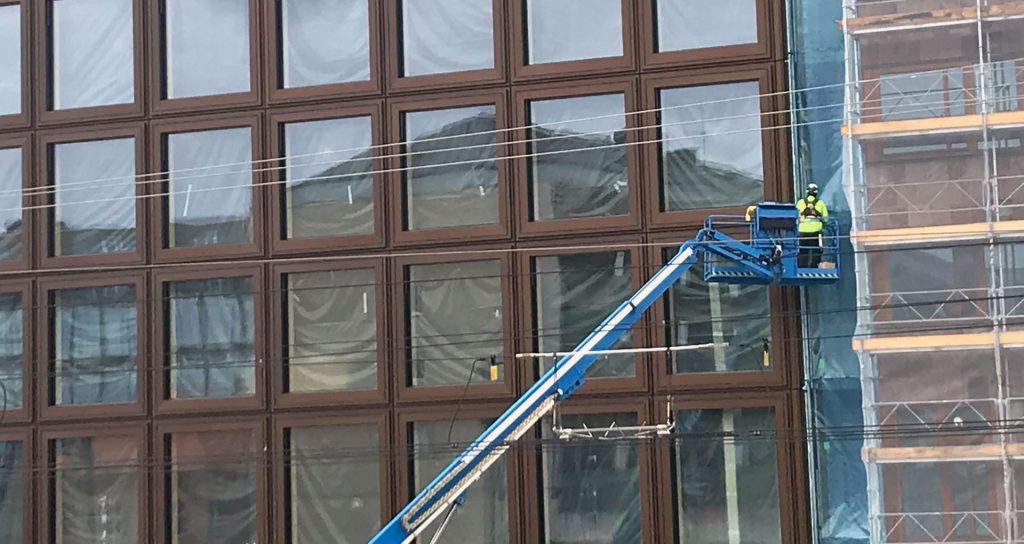 Bilden visar ett pågående bygge av ett stort hus med glasfönster och en byggarbetare på en kran. I fönsterrutorna speglas ett bostadshus.